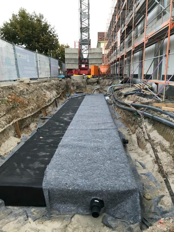 Regenwasserrückhalte-Anlage Baugrube mit schwarzer Struktur auf einem unterirdischen Fundament und Baugeräten im Hintergrund.