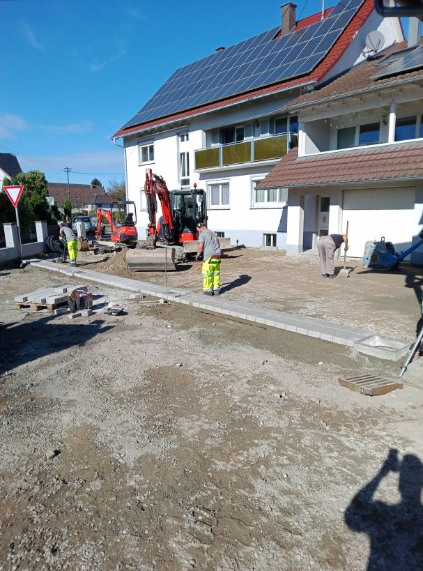 Anlegen der Entwässerungswege Bauarbeiten mit Bagger und Arbeiter auf einem unbefestigten Gelände neben einem Mehrfamilienhaus.