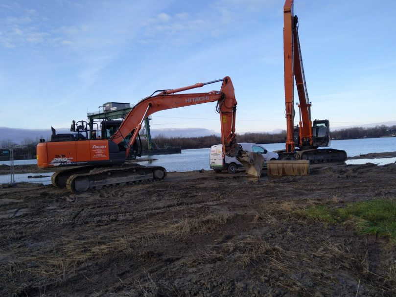Baufeldvorbereitung Bagger arbeiten am Ufer eines Gewässers, umgeben von Erde und Gras.