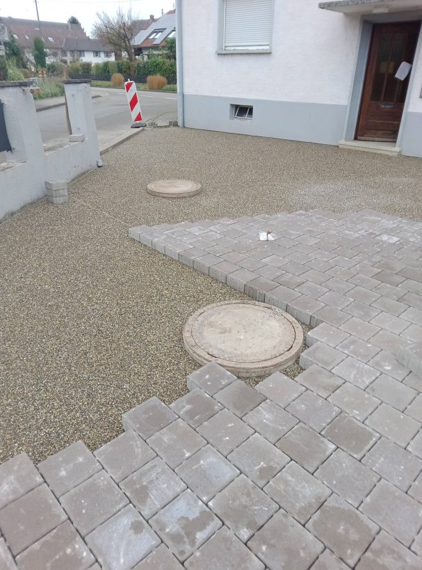 Pflasterarbeiten Kiesfläche mit zwei runden Kanalschächten und Pflastersteinen.