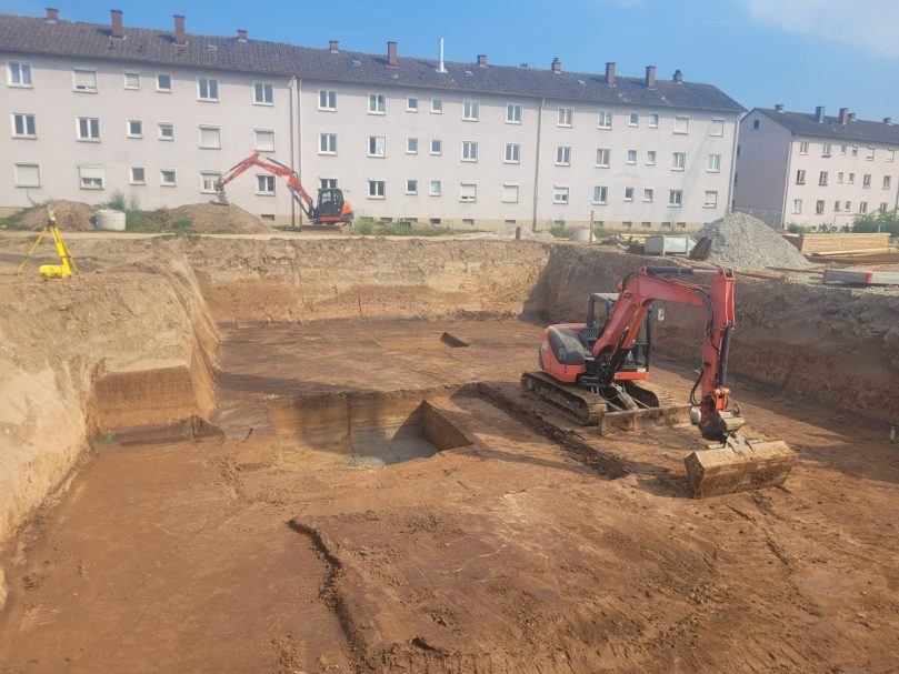 Baugrubenaushub Bagger arbeitet auf einer Baustelle mit gegrabenem Boden und Wohngebäuden im Hintergrund.