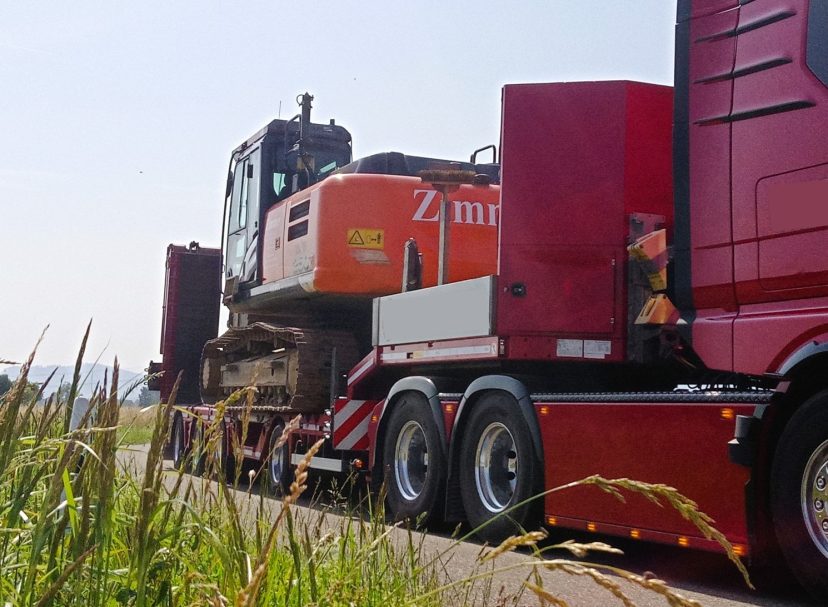 Baumaschinentransport Lkw mit Baugerät auf einer Straße, umgeben von hohem Gras.