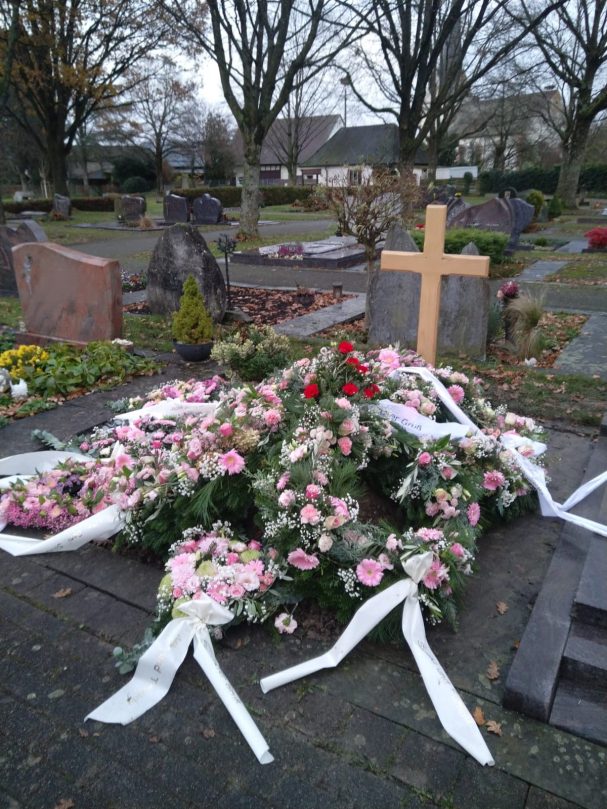 Abgedecktes Grab Blumengestecke mit weißen Bändern auf einem Friedhof, neben einem Kreuz aus Holz.