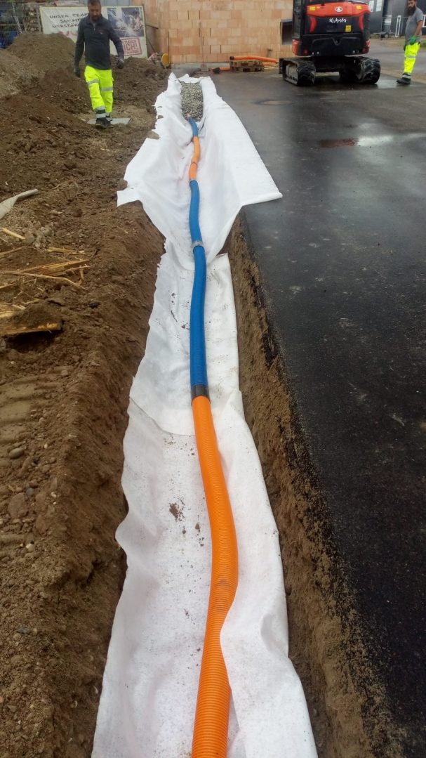 Drainageleitung im Graben Graben mit einer blauen und einer orangefarbenen Leitung, umgeben von Erde.